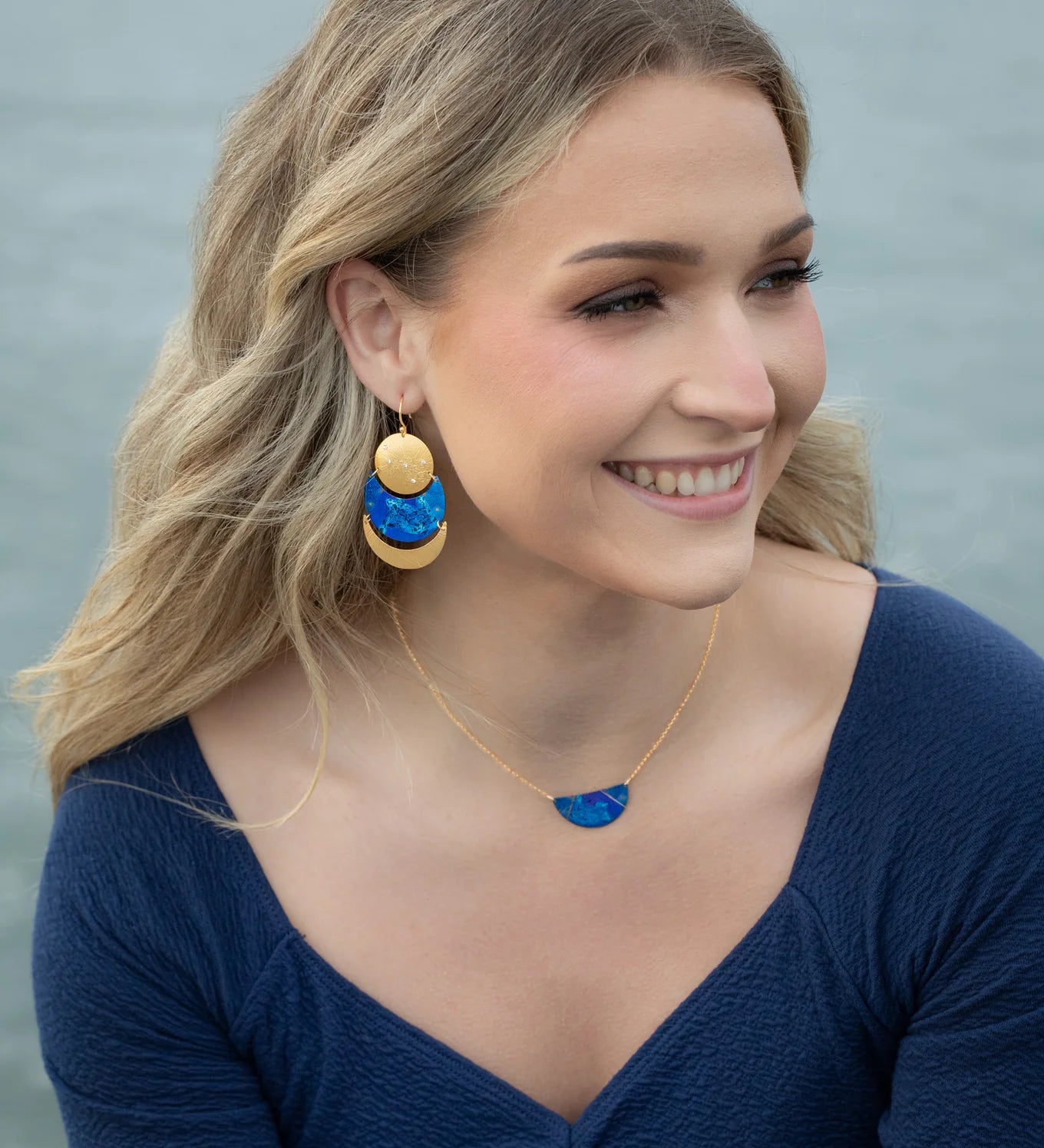 A smiling woman with wavy blonde hair wears a midnight blue top, blue and gold statement earrings, and the Holly Yashi Orion Pendant/ Midnight Blue Necklace, seated outdoors near water.
