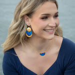 A smiling woman with wavy blonde hair wears a midnight blue top, blue and gold statement earrings, and the Holly Yashi Orion Pendant/ Midnight Blue Necklace, seated outdoors near water.