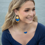 A smiling woman with wavy blonde hair wears a midnight blue top, blue and gold statement earrings, and the Holly Yashi Orion Pendant/ Midnight Blue Necklace, seated outdoors near water.