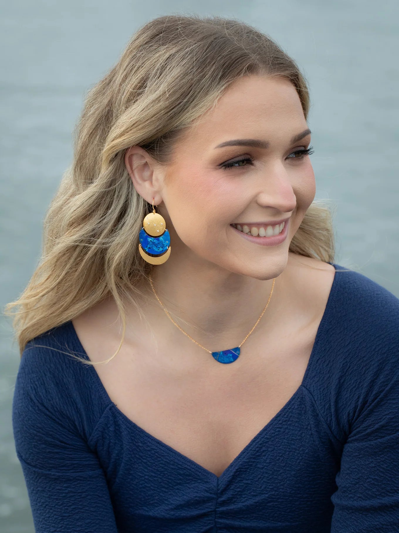 A smiling woman with wavy blonde hair wears a midnight blue top, blue and gold statement earrings, and the Holly Yashi Orion Pendant/ Midnight Blue Necklace, seated outdoors near water.