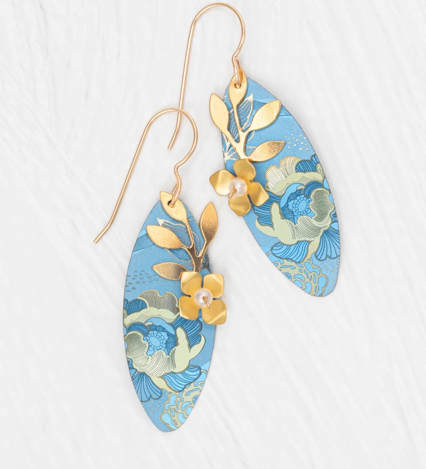 The Alyssa Sky Blue Earrings by Holly Yashi feature elongated blue ovals with gold floral patterns, leaf and flower accents, and hand-colored niobium, shown on a white textured background.