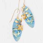 The Alyssa Sky Blue Earrings by Holly Yashi feature elongated blue ovals with gold floral patterns, leaf and flower accents, and hand-colored niobium, shown on a white textured background.