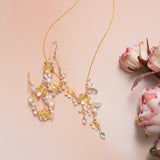 The Rafaela Gold & White Necklace by Holly Yashi, featuring floral and leaf motifs with pearls and crystals, is displayed on a soft pink surface beside pale pink roses.