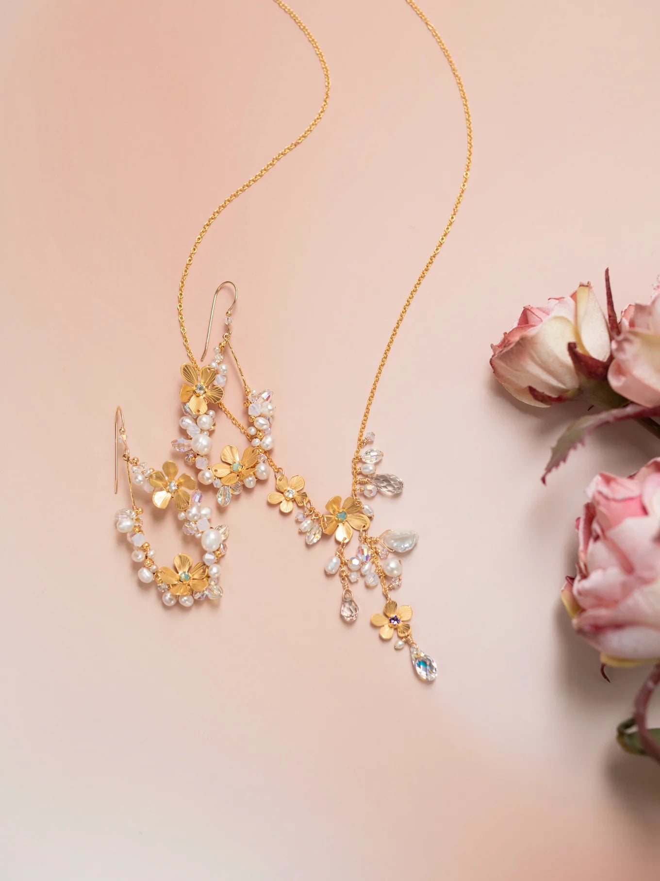 The Rafaela Gold & White Necklace by Holly Yashi, featuring floral and leaf motifs with pearls and crystals, is displayed on a soft pink surface beside pale pink roses.
