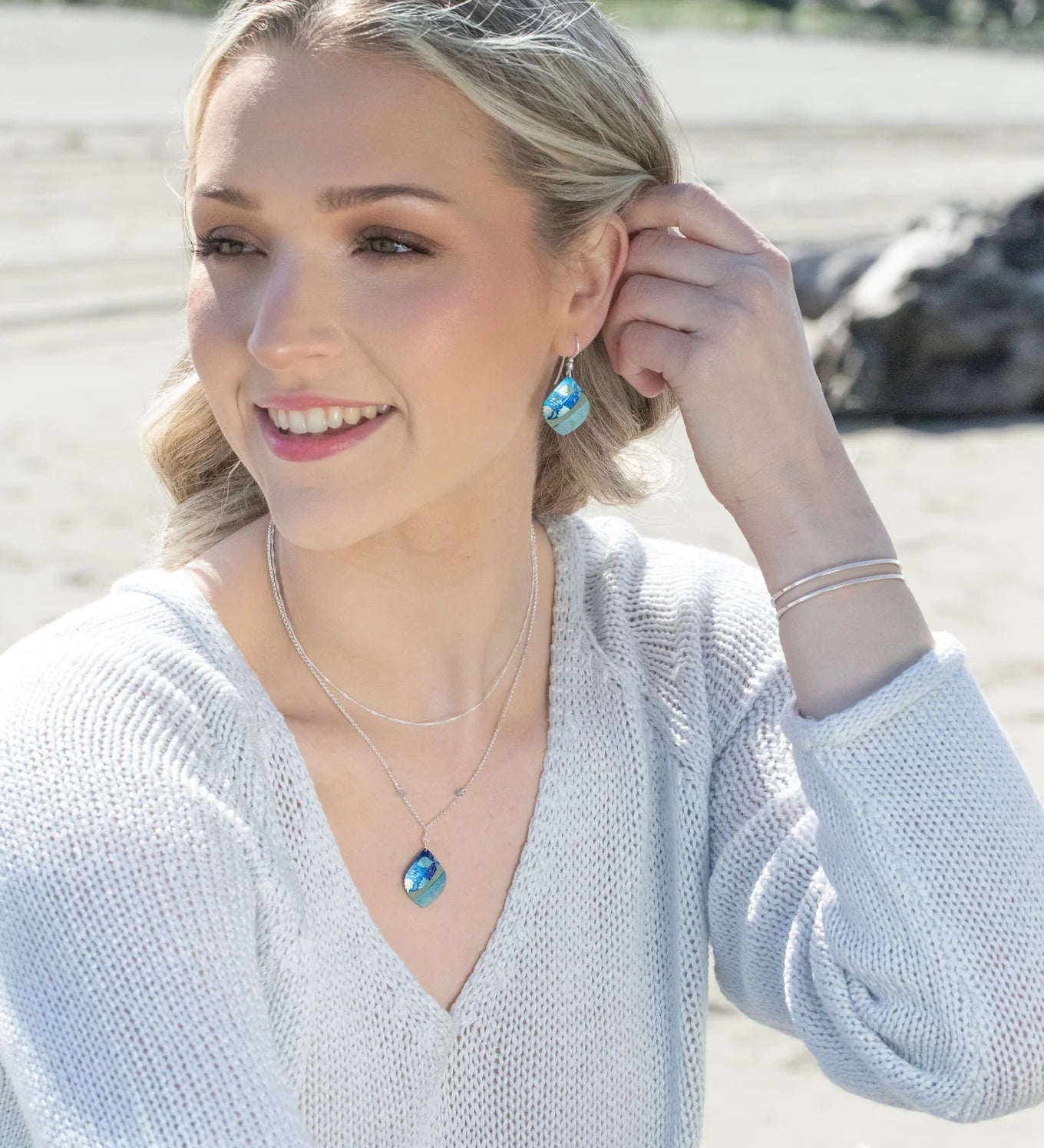 A blonde woman smiles on a sandy beach, wearing a white sweater, silver jewelry, and Lani Kai Blue Earrings by Holly Yashi. She touches her hypoallergenic earring with rocks and water in the background.