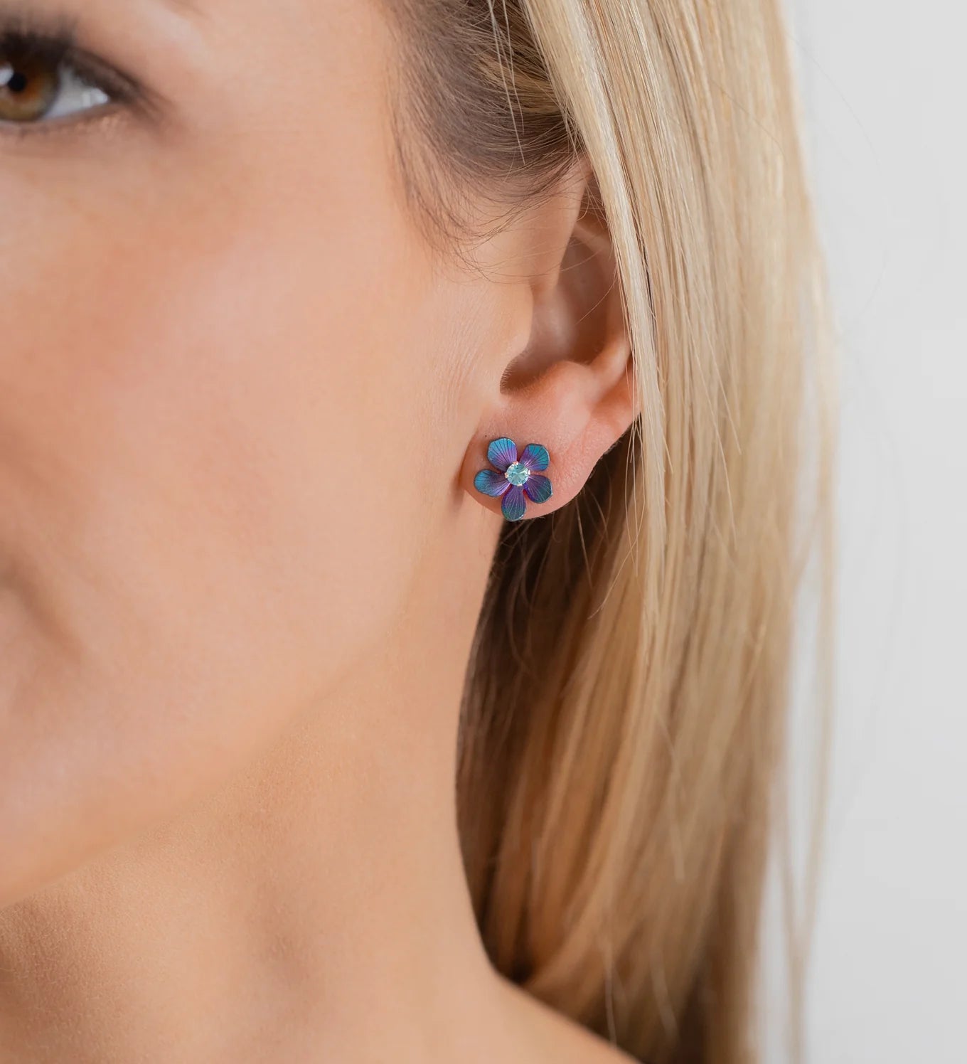 Close-up of a woman wearing Holly Yashi's Petite Plumeria Post Gold/Champagne Earrings—small, flower-shaped studs with gold tones. She has straight blonde hair and light skin against a plain background. Perfect for garden parties.