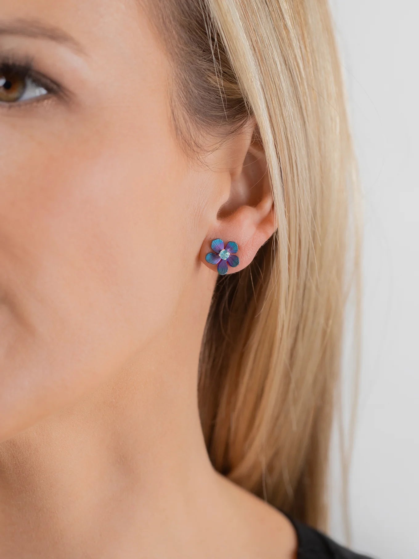 Close-up of a woman wearing Holly Yashi's Petite Plumeria Post Gold/Champagne Earrings—small, flower-shaped studs with gold tones. She has straight blonde hair and light skin against a plain background. Perfect for garden parties.