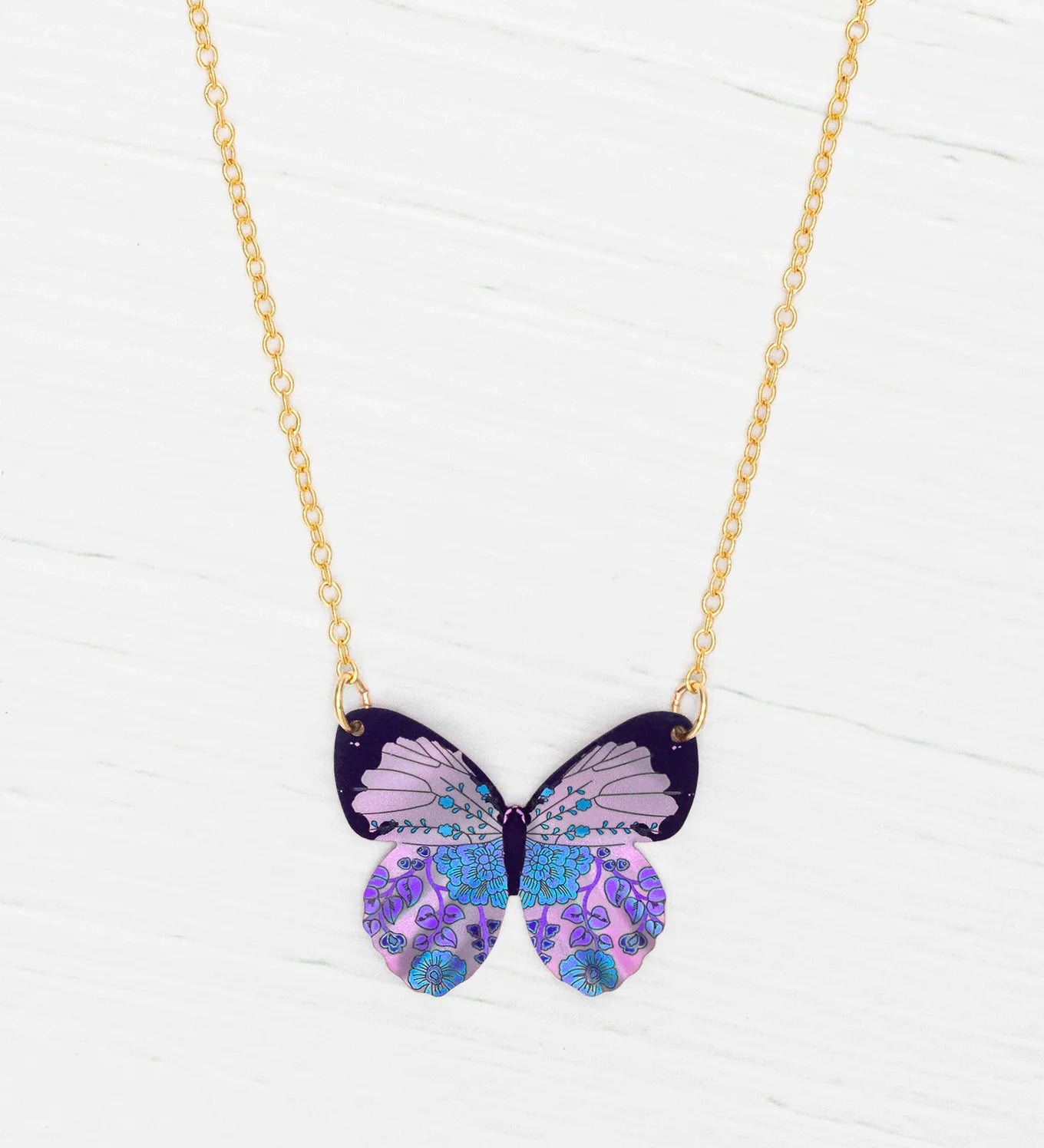 The Blooming Butterfly Radiant Purple Necklace by Holly Yashi features an iridescent niobium butterfly pendant with purple, blue, and lavender patterns on a gold adjustable chain, shown on a white wooden surface.