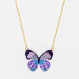 The Blooming Butterfly Radiant Purple Necklace by Holly Yashi features an iridescent niobium butterfly pendant with purple, blue, and lavender patterns on a gold adjustable chain, shown on a white wooden surface.