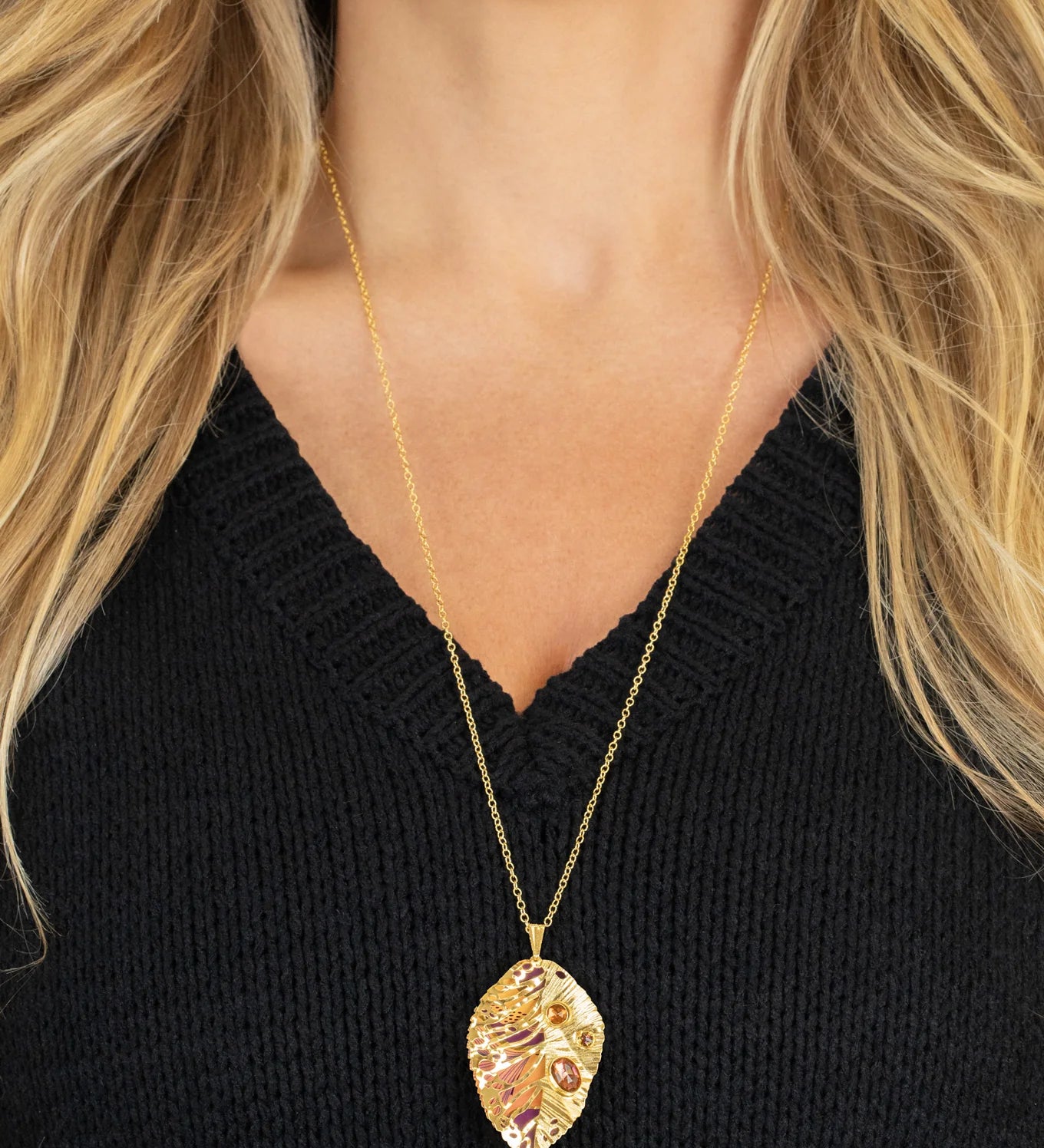 A woman wears the Perennial Elm Indigo Necklace by Holly Yashi, featuring a large, intricately designed leaf-shaped pendant on an adjustable gold chain, styled with her long blonde hair and a black V-neck sweater.