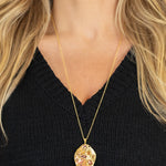 A woman wears the Perennial Elm Indigo Necklace by Holly Yashi, featuring a large, intricately designed leaf-shaped pendant on an adjustable gold chain, styled with her long blonde hair and a black V-neck sweater.