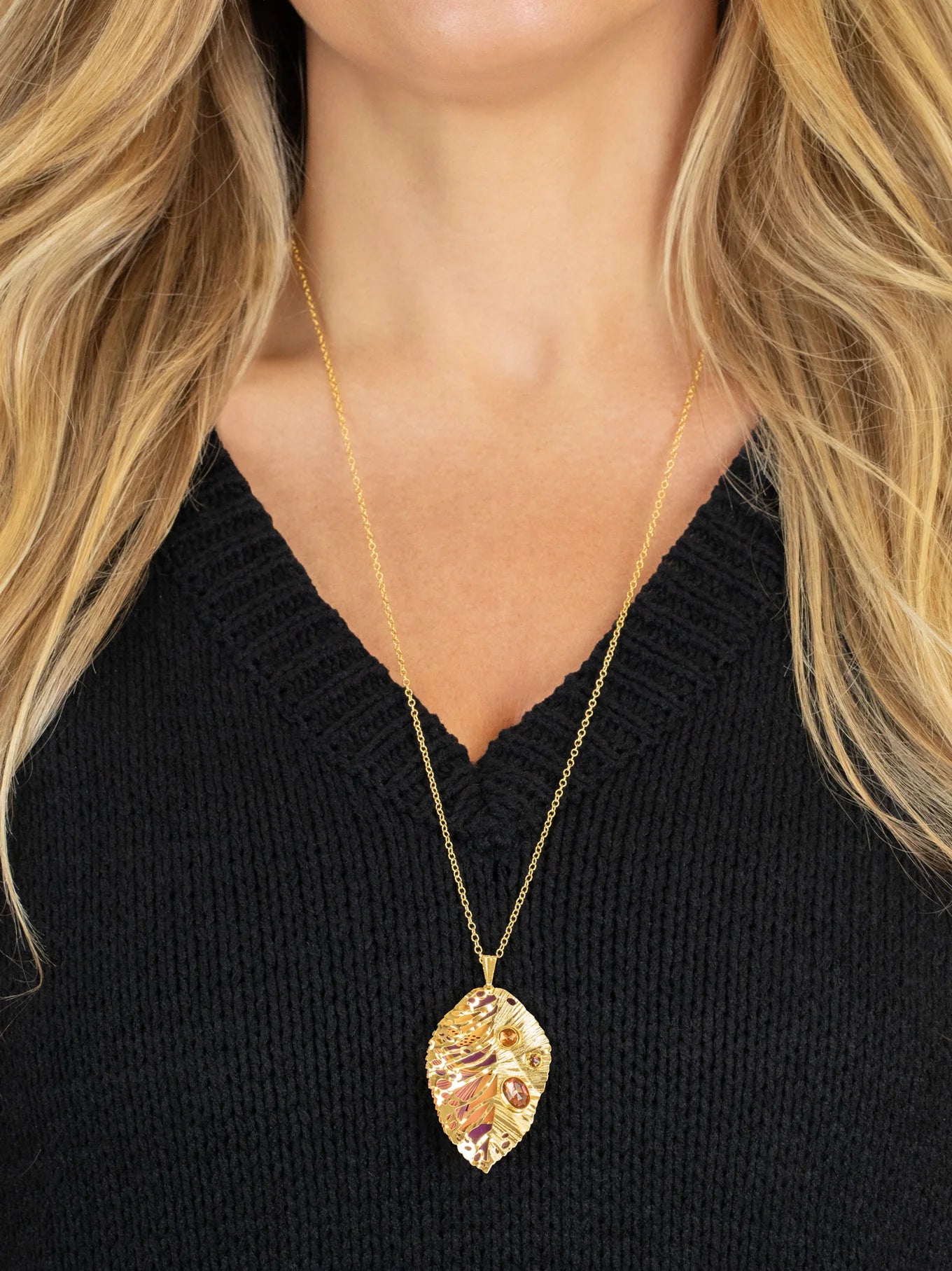A woman wears the Perennial Elm Indigo Necklace by Holly Yashi, featuring a large, intricately designed leaf-shaped pendant on an adjustable gold chain, styled with her long blonde hair and a black V-neck sweater.