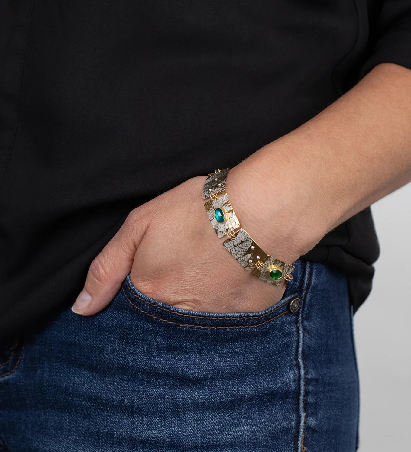A person in blue jeans and a black shirt has their hand in their pocket, showcasing the Holly Yashi Gilded Petra Emerald Bracelet adorned with emerald green crystals and shimmering blue gemstones.