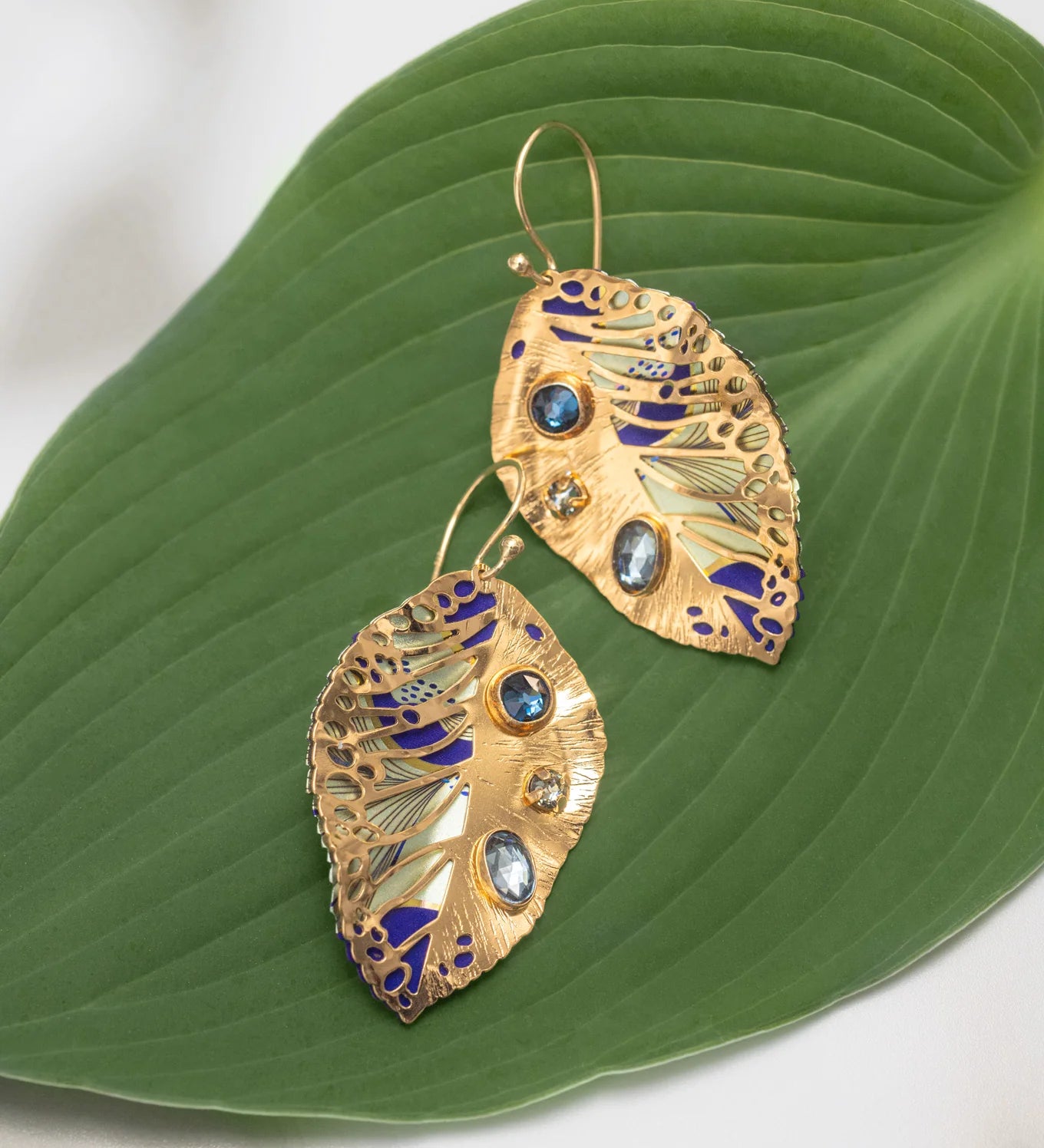 The Perennial Elm Indigo Earrings by Holly Yashi feature a leaf-shaped design with blue and clear stones plus purple enamel accents, beautifully displayed on a large green leaf background.