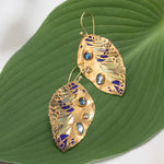 The Perennial Elm Indigo Earrings by Holly Yashi feature a leaf-shaped design with blue and clear stones plus purple enamel accents, beautifully displayed on a large green leaf background.
