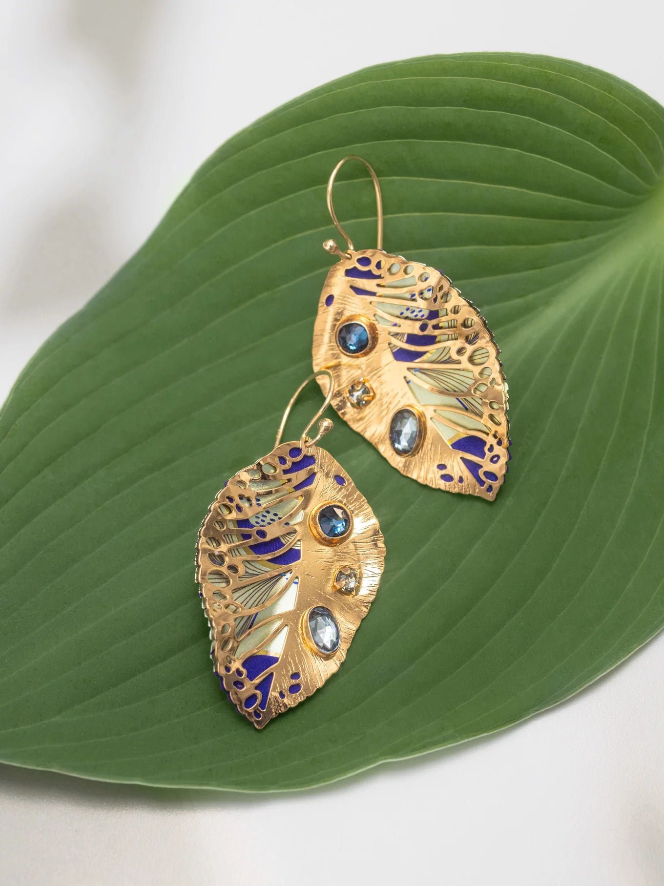 The Perennial Elm Indigo Earrings by Holly Yashi feature a leaf-shaped design with blue and clear stones plus purple enamel accents, beautifully displayed on a large green leaf background.