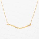 The Holly Yashi Zara Bar Gold Necklace features a slender, curved gold bar with clear gemstones. This hypoallergenic, minimal design is an elegant statement piece for any occasion.