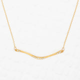 The Holly Yashi Zara Bar Gold Necklace features a slender, curved gold bar with clear gemstones. This hypoallergenic, minimal design is an elegant statement piece for any occasion.