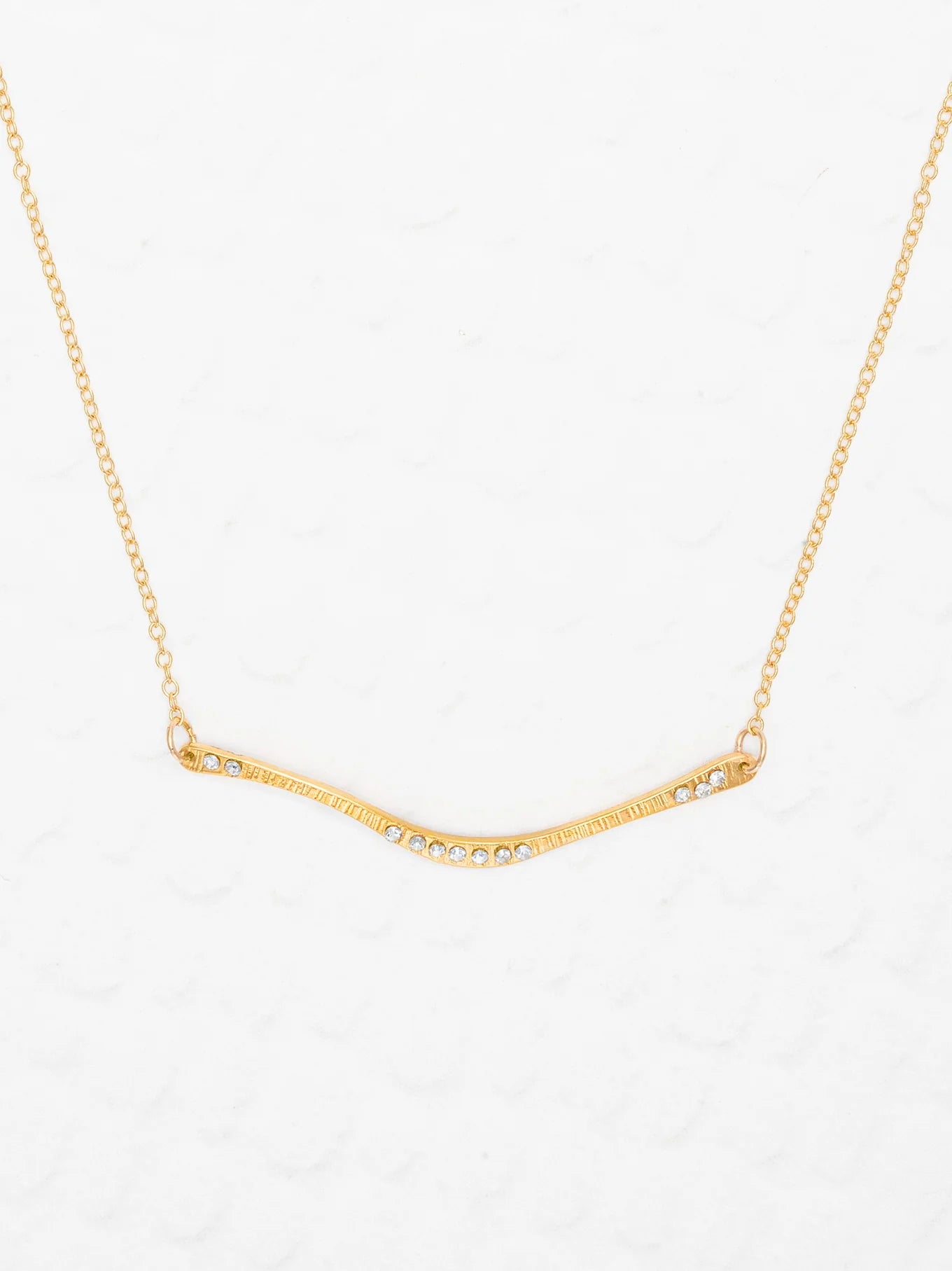 The Holly Yashi Zara Bar Gold Necklace features a slender, curved gold bar with clear gemstones. This hypoallergenic, minimal design is an elegant statement piece for any occasion.
