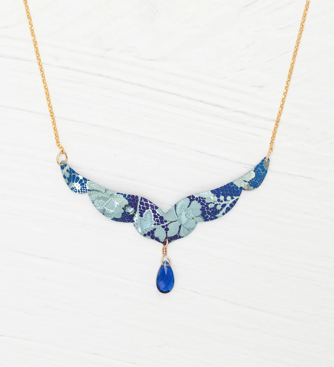 The Isla Forever Blue Necklace by Holly Yashi features a gold chain with a turquoise, wing-shaped niobium pendant decorated with a floral pattern and a central blue teardrop gemstone, shown on a white textured background.
