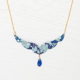 The Isla Forever Blue Necklace by Holly Yashi features a gold chain with a turquoise, wing-shaped niobium pendant decorated with a floral pattern and a central blue teardrop gemstone, shown on a white textured background.