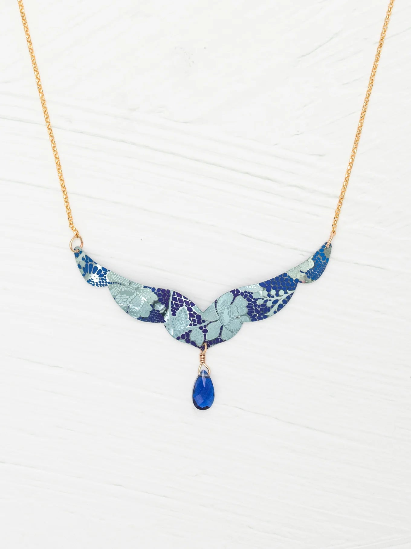 The Isla Forever Blue Necklace by Holly Yashi features a gold chain with a turquoise, wing-shaped niobium pendant decorated with a floral pattern and a central blue teardrop gemstone, shown on a white textured background.