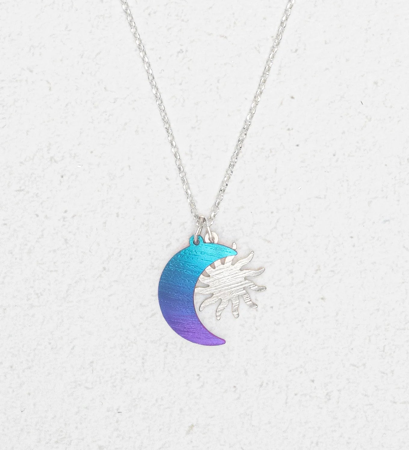 The Sun and Moon Calypso Necklace by Holly Yashi features a silver sun overlapping a crescent moon in a blue to purple gradient, displayed on a white textured background—ideal for celestial jewelry lovers.