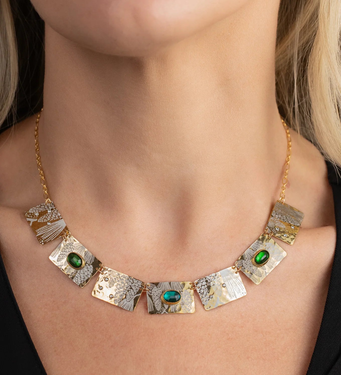 A close-up of a woman's neck wearing the Gilded Petra Emerald Necklace by Holly Yashi—a gold chain with textured rectangular plates and three emerald green crystals—paired with a black top for modern Victorian flair.