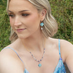 A woman with short blonde hair wears Holly Yashi’s Elena Violet Earrings, a light blue floral dress, and a necklace. She stands outdoors amid greenery, looking to the side with a slight smile.