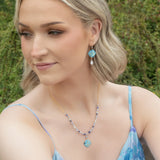 A woman with short blonde hair wears Holly Yashi’s Elena Violet Earrings, a light blue floral dress, and a necklace. She stands outdoors amid greenery, looking to the side with a slight smile.