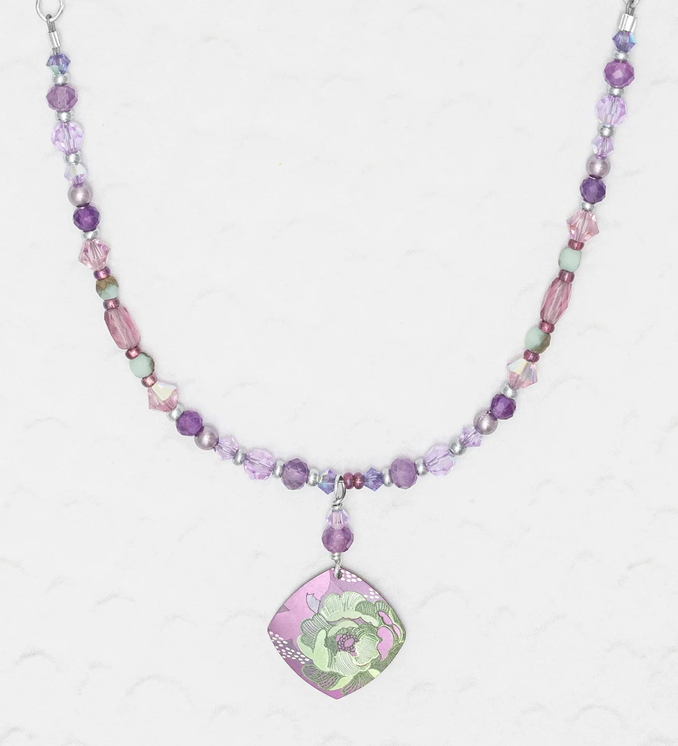 The Elena Beaded Necklace Violet by Holly Yashi features purple, pink, and green beads with a square floral green and purple pendant, shown on a textured white background.