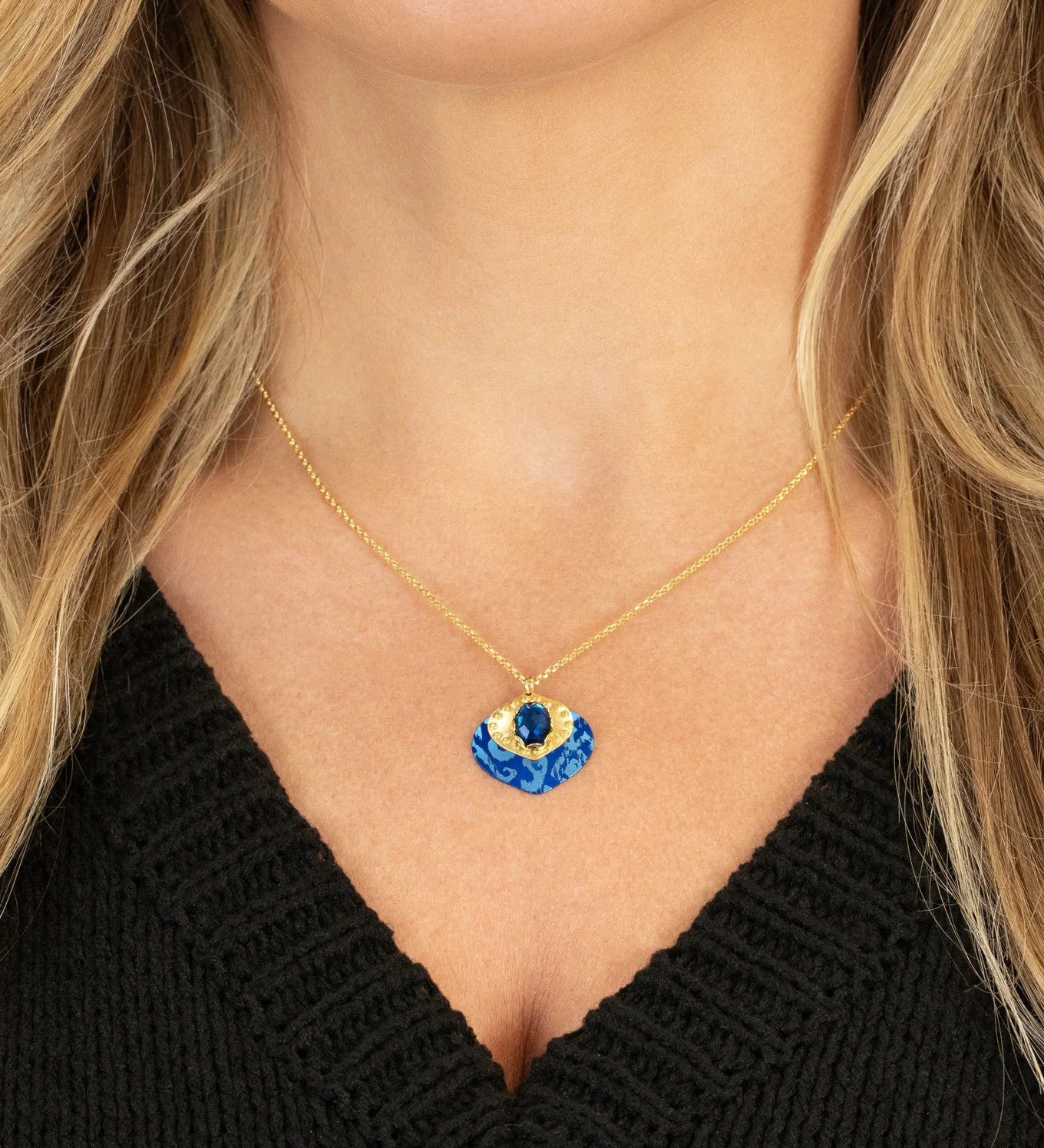 A woman wears the Holly Yashi Arabella Sapphire Blue Necklace, featuring an adjustable gold chain and a vibrant blue and gold glass stone pendant. She pairs it with a black V-neck top.