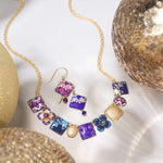 A gold chain necklace and Holly Yashi’s Garden Soiree Vintage Burgundy Earrings—square purple, pink, and blue pendants with intricate gold designs—are displayed among glittery metallic spheres on a white surface.