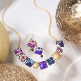 A gold chain necklace and Holly Yashi’s Garden Soiree Vintage Burgundy Earrings—square purple, pink, and blue pendants with intricate gold designs—are displayed among glittery metallic spheres on a white surface.
