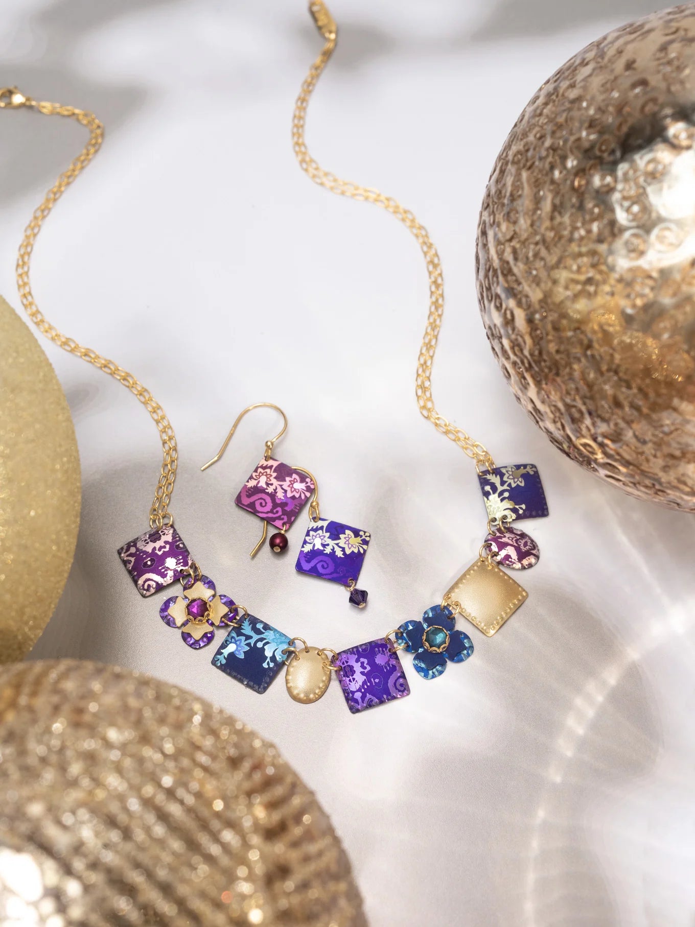 A gold chain necklace and Holly Yashi’s Garden Soiree Vintage Burgundy Earrings—square purple, pink, and blue pendants with intricate gold designs—are displayed among glittery metallic spheres on a white surface.