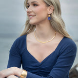 A woman with long blonde hair, in a navy blue top and gold jewelry, sits outdoors by the water, smiling and wearing Holly Yashi’s Danica Earrings in Midnight Blue.