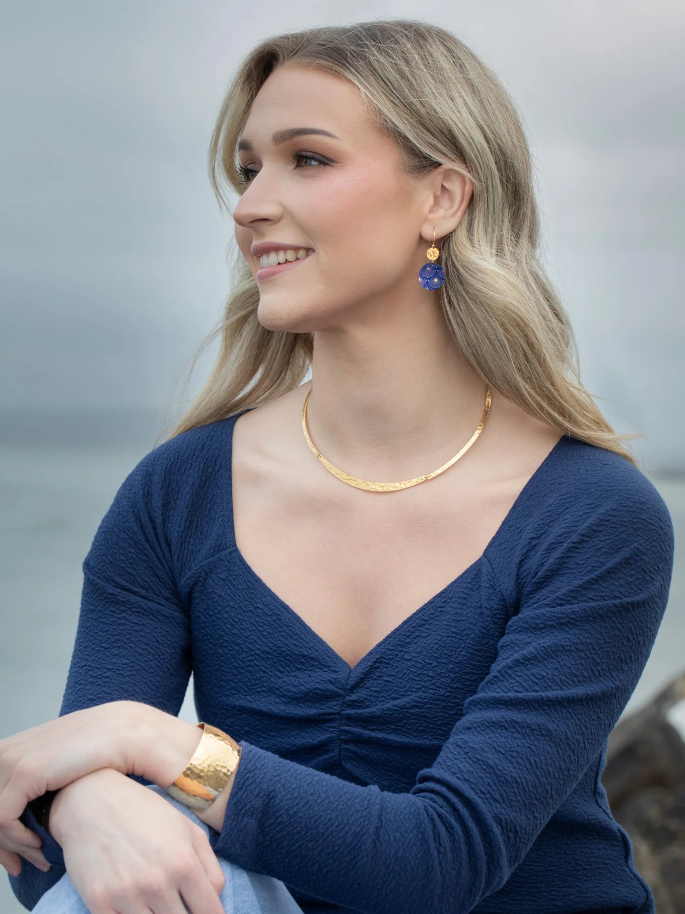 A woman with long blonde hair, in a navy blue top and gold jewelry, sits outdoors by the water, smiling and wearing Holly Yashi’s Danica Earrings in Midnight Blue.