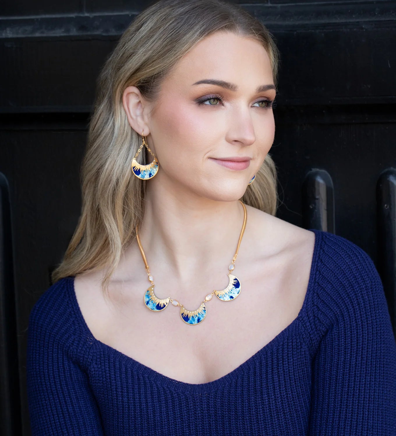 A woman with long blonde hair, wearing a blue sweater and Holly Yashis Selena/Midnight Blue earrings and matching necklace, sits before a black background, gazing slightly to the side with a gentle smile.