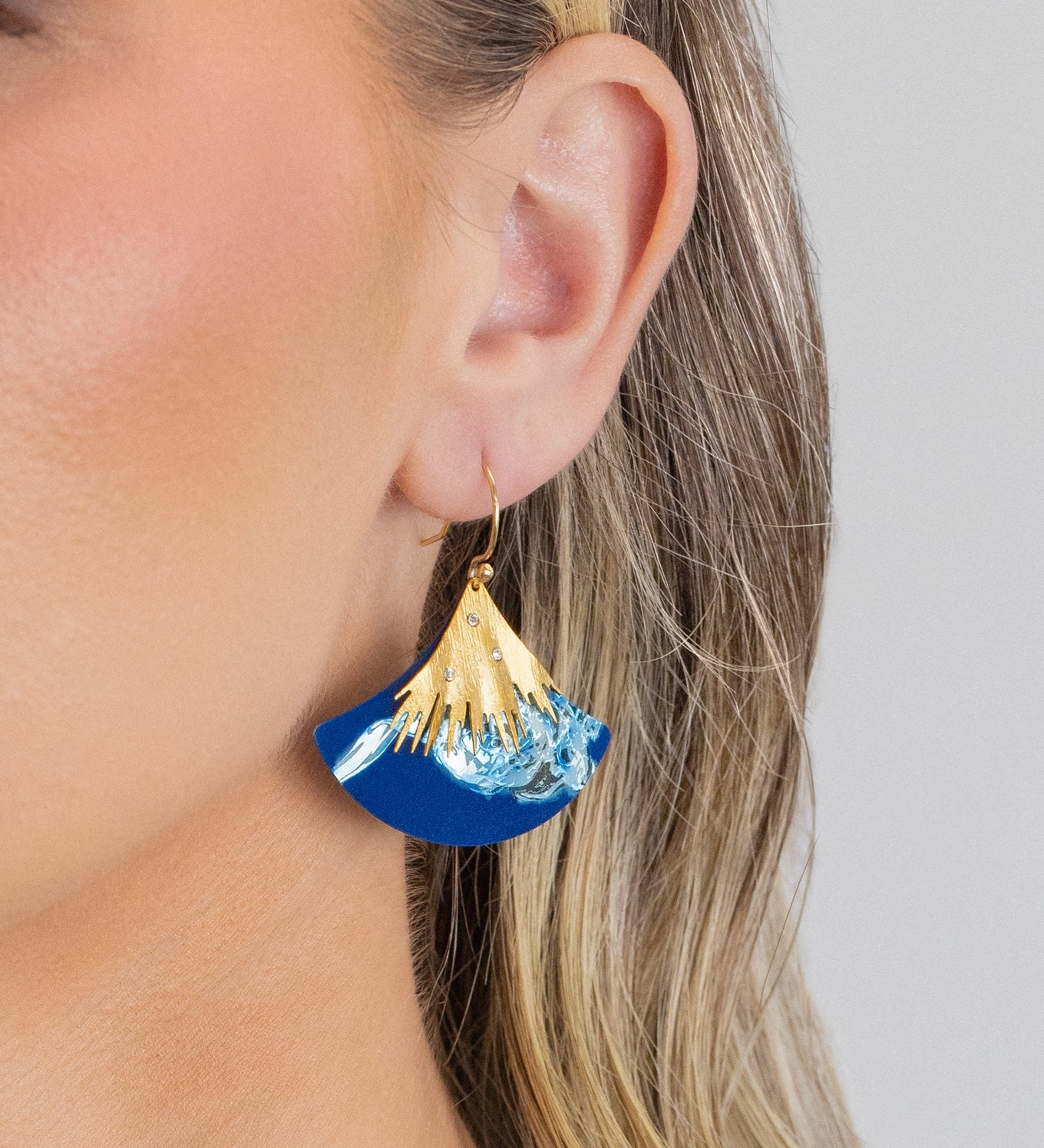 A close-up shows a woman with blonde hair tucked behind her ear, wearing the Holly Yashi Aster Earrings in Midnight Blue—hypoallergenic statement earrings with a gold, fan-shaped top and wave-like bottom.