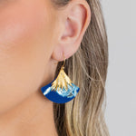 A close-up shows a woman with blonde hair tucked behind her ear, wearing the Holly Yashi Aster Earrings in Midnight Blue—hypoallergenic statement earrings with a gold, fan-shaped top and wave-like bottom.