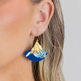 A close-up shows a woman with blonde hair tucked behind her ear, wearing the Holly Yashi Aster Earrings in Midnight Blue—hypoallergenic statement earrings with a gold, fan-shaped top and wave-like bottom.