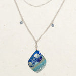 The Lani/Kai Blue Necklace by Holly Yashi features layered silver with a large teardrop pendant in blue and teal leaf patterns, plus two square blue charms. This hypoallergenic piece adds a seaside touch to any look.