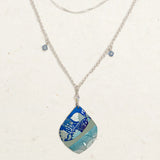 The Lani/Kai Blue Necklace by Holly Yashi features layered silver with a large teardrop pendant in blue and teal leaf patterns, plus two square blue charms. This hypoallergenic piece adds a seaside touch to any look.