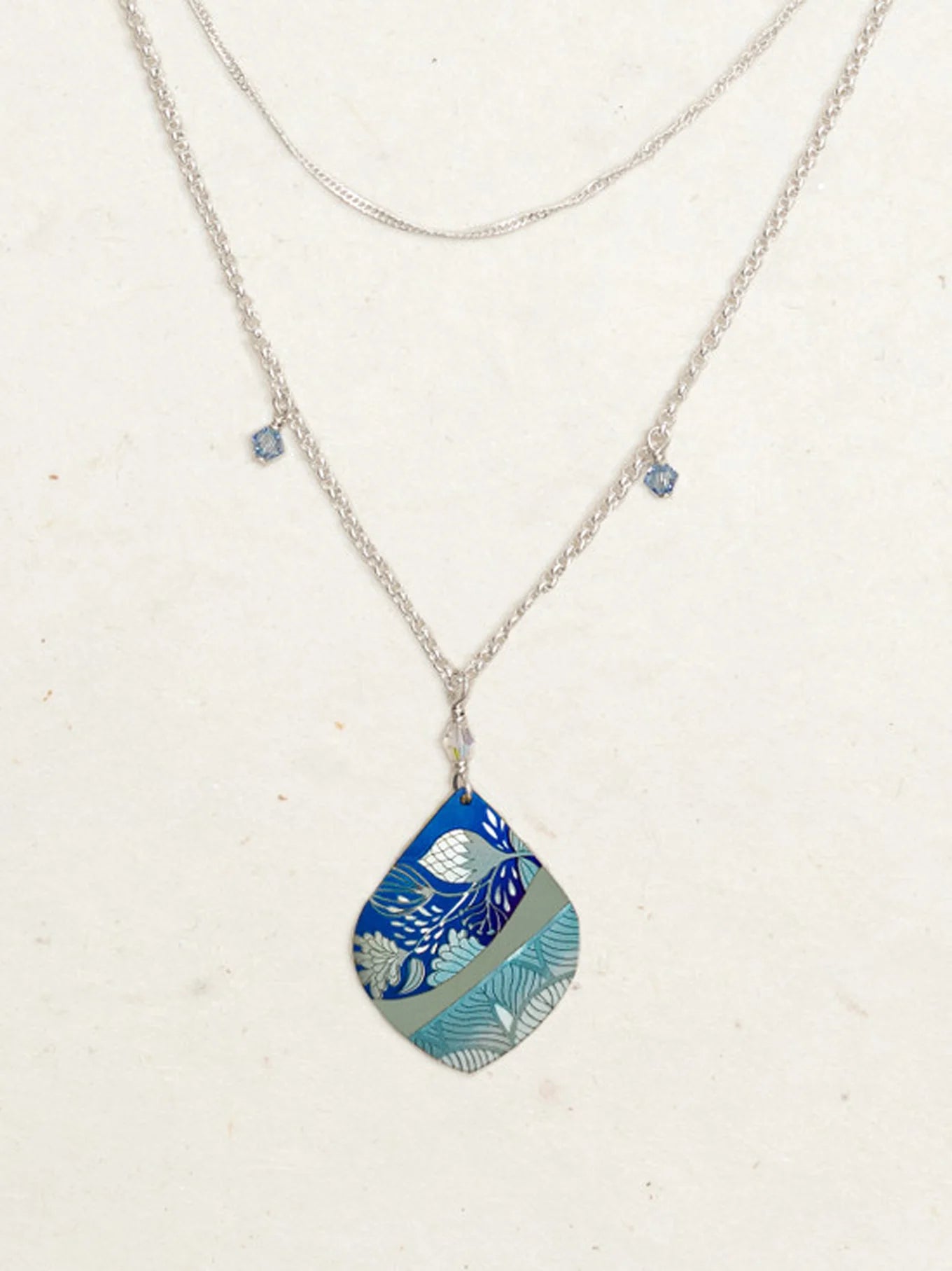 The Lani/Kai Blue Necklace by Holly Yashi features layered silver with a large teardrop pendant in blue and teal leaf patterns, plus two square blue charms. This hypoallergenic piece adds a seaside touch to any look.