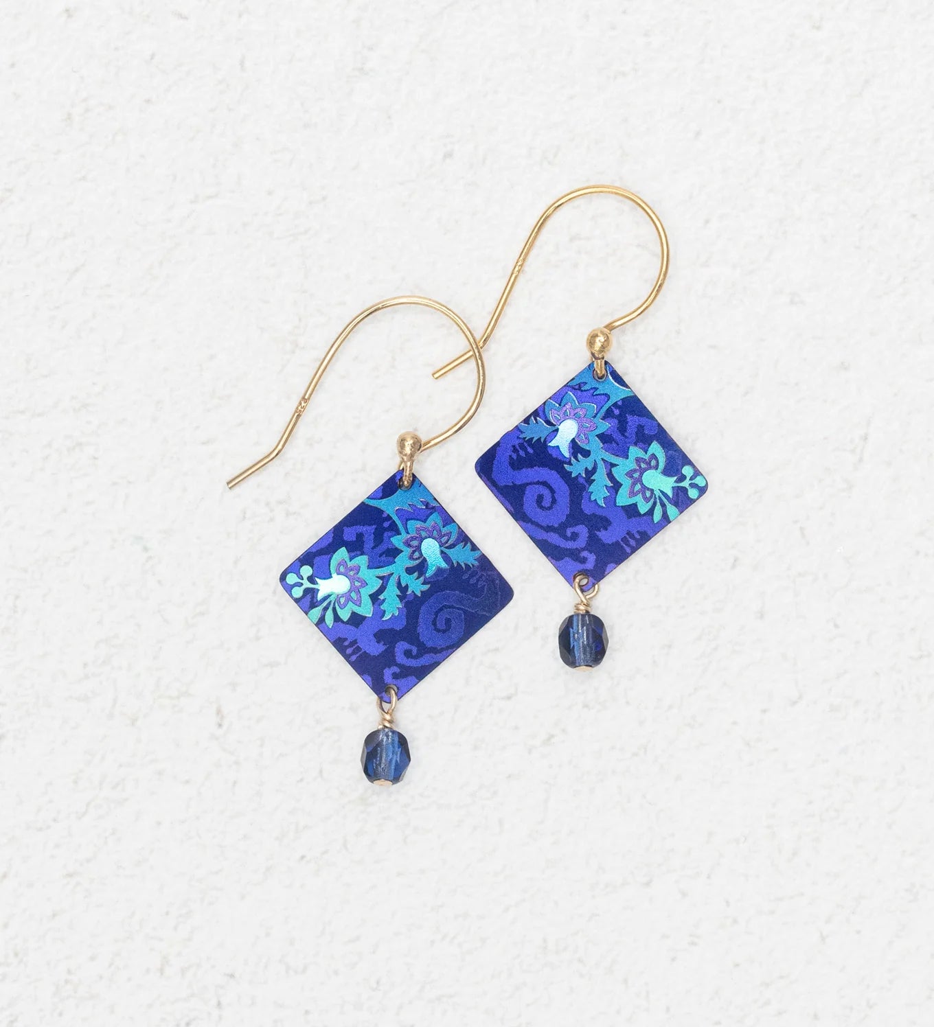 The Holly Yashi Garden Soiree Sapphire Blue Earrings are gold hook, hypoallergenic earrings with sapphire blue square floral pendants and a dangling blue bead, shown on a light textured background.