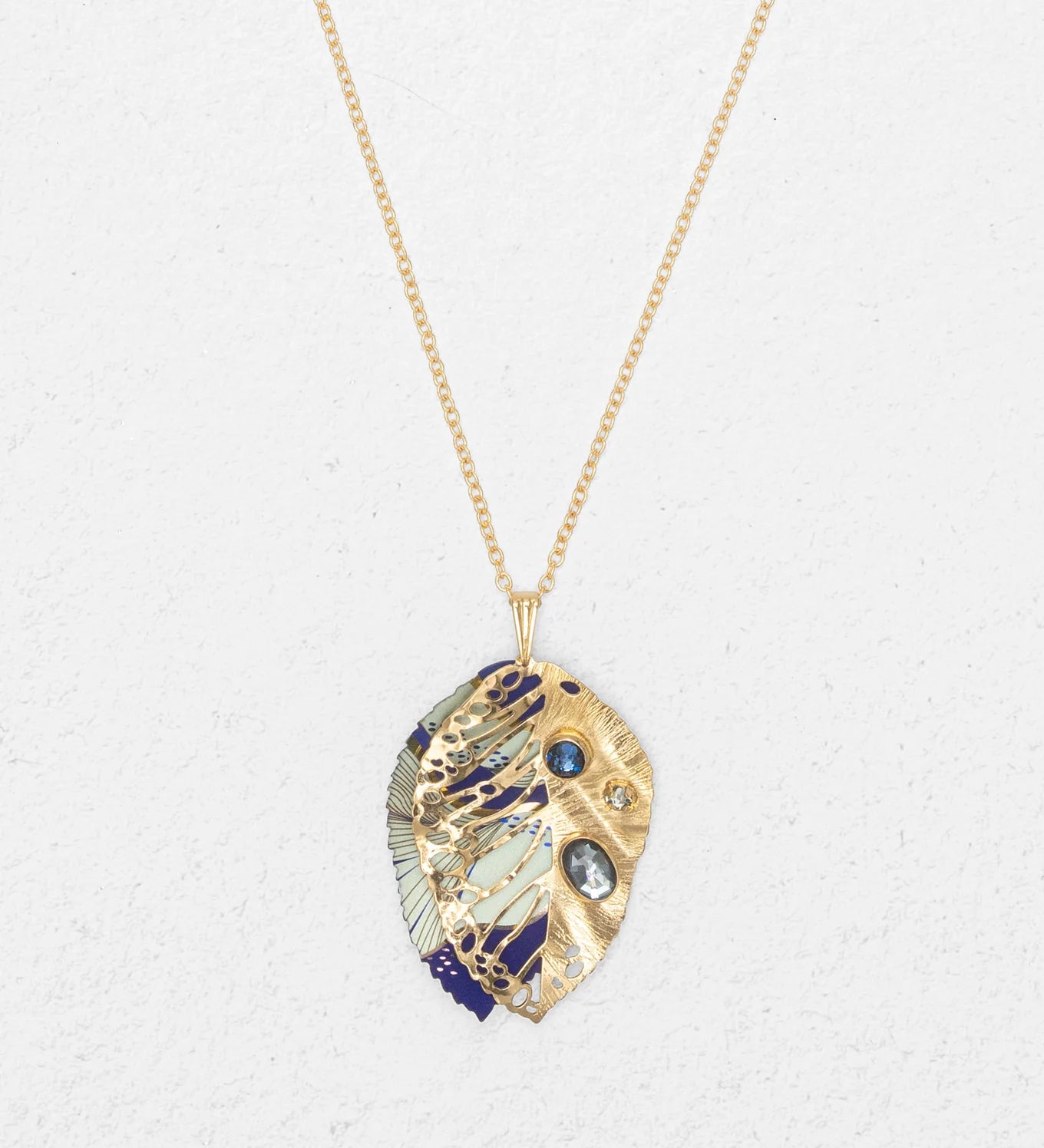 The Perennial Elm Indigo Necklace by Holly Yashi features a gold adjustable chain with a leaf-shaped gemstone pendant, intricate cut-out patterns, blue and white enamel details, and blue gemstone accents, displayed on a light textured background.
