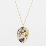 The Perennial Elm Indigo Necklace by Holly Yashi features a gold adjustable chain with a leaf-shaped gemstone pendant, intricate cut-out patterns, blue and white enamel details, and blue gemstone accents, displayed on a light textured background.