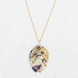 The Perennial Elm Indigo Necklace by Holly Yashi features a gold adjustable chain with a leaf-shaped gemstone pendant, intricate cut-out patterns, blue and white enamel details, and blue gemstone accents, displayed on a light textured background.