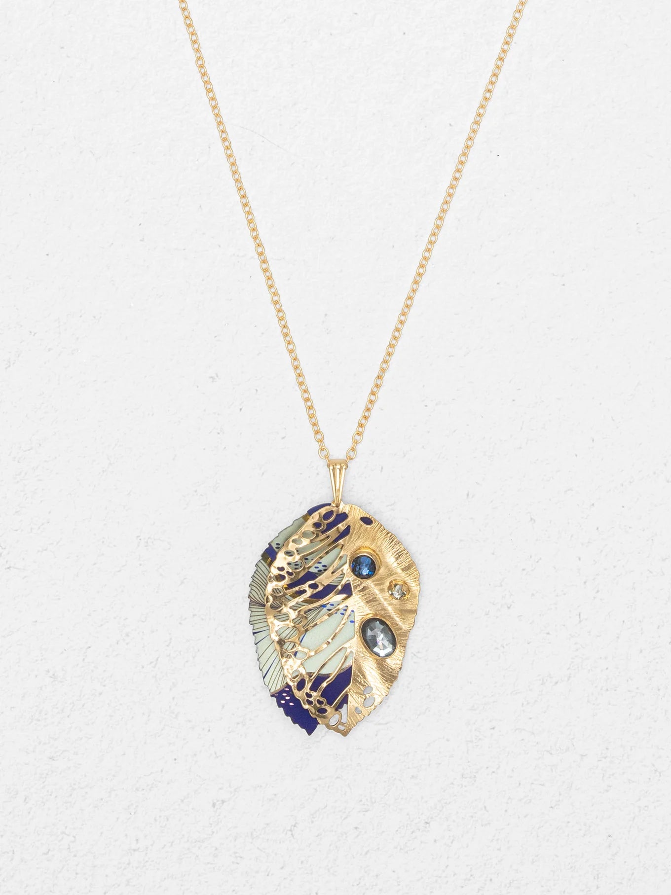The Perennial Elm Indigo Necklace by Holly Yashi features a gold adjustable chain with a leaf-shaped gemstone pendant, intricate cut-out patterns, blue and white enamel details, and blue gemstone accents, displayed on a light textured background.