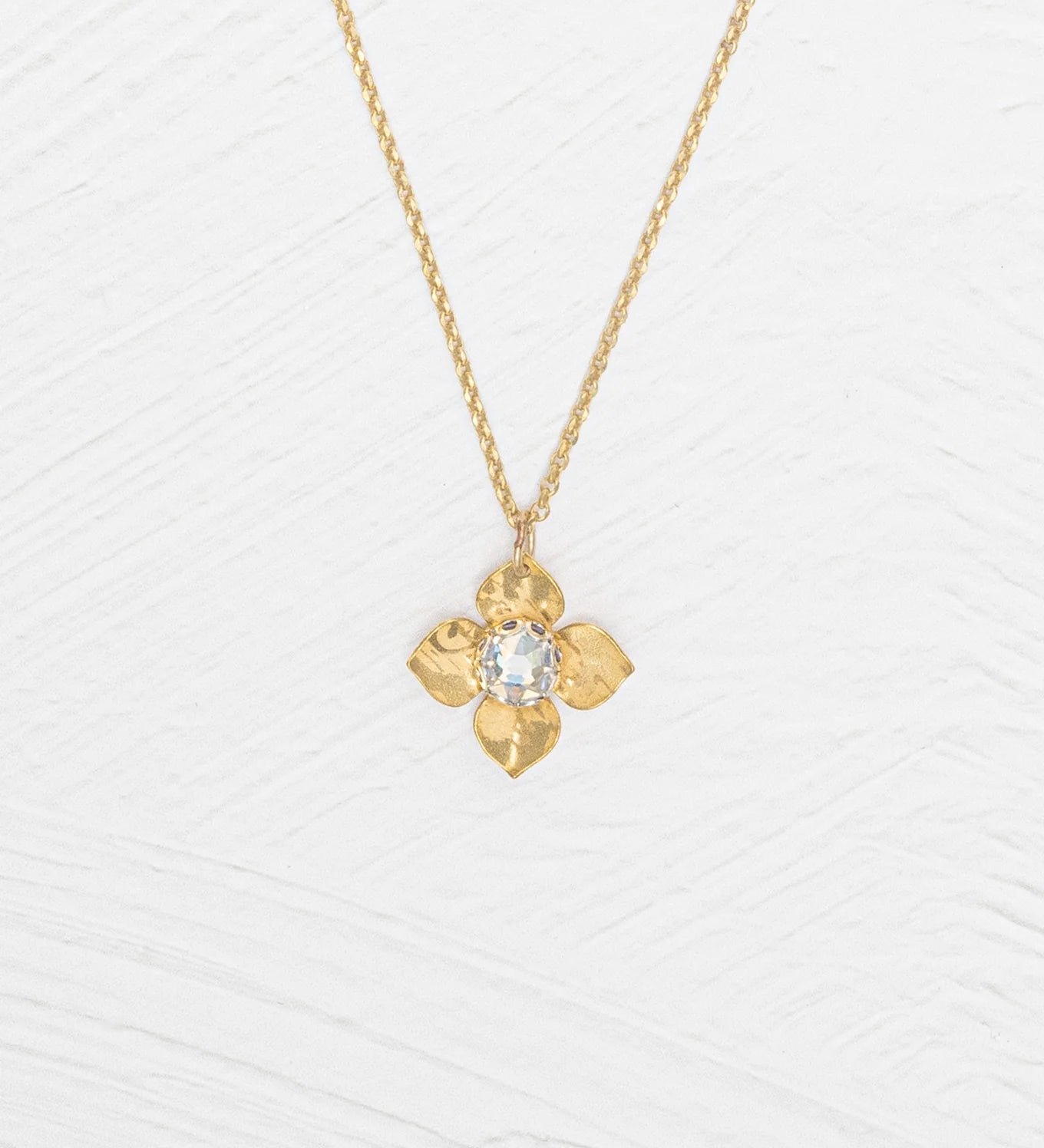 The Holly Yashi Petite Fleur Gold Necklace showcases a four-petal flower pendant with a sparkling Austrian crystal at the center, elegantly set against a white textured surface.
